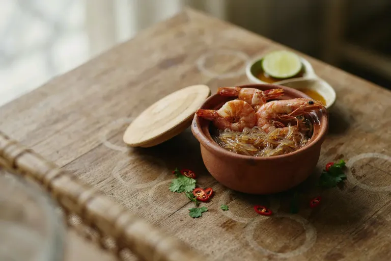 A small clay pot of glass noodles with whole prawns, lid set to one side