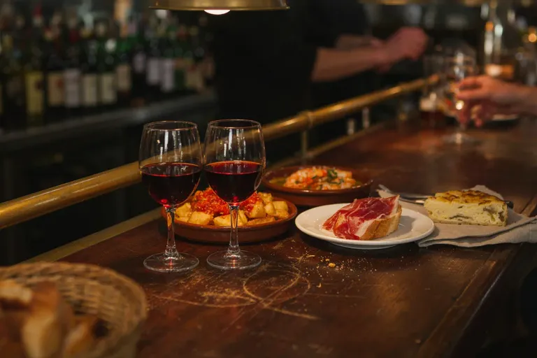 A bar counter with glasses of red wine, a plate of sliced ham and a wedge of Spanish omelette