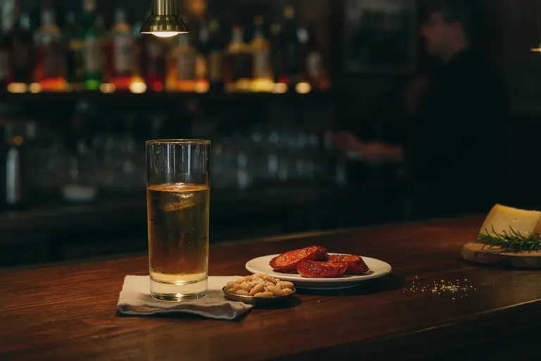 A glass of beer and a plate of chorizo on a dark wooden bar